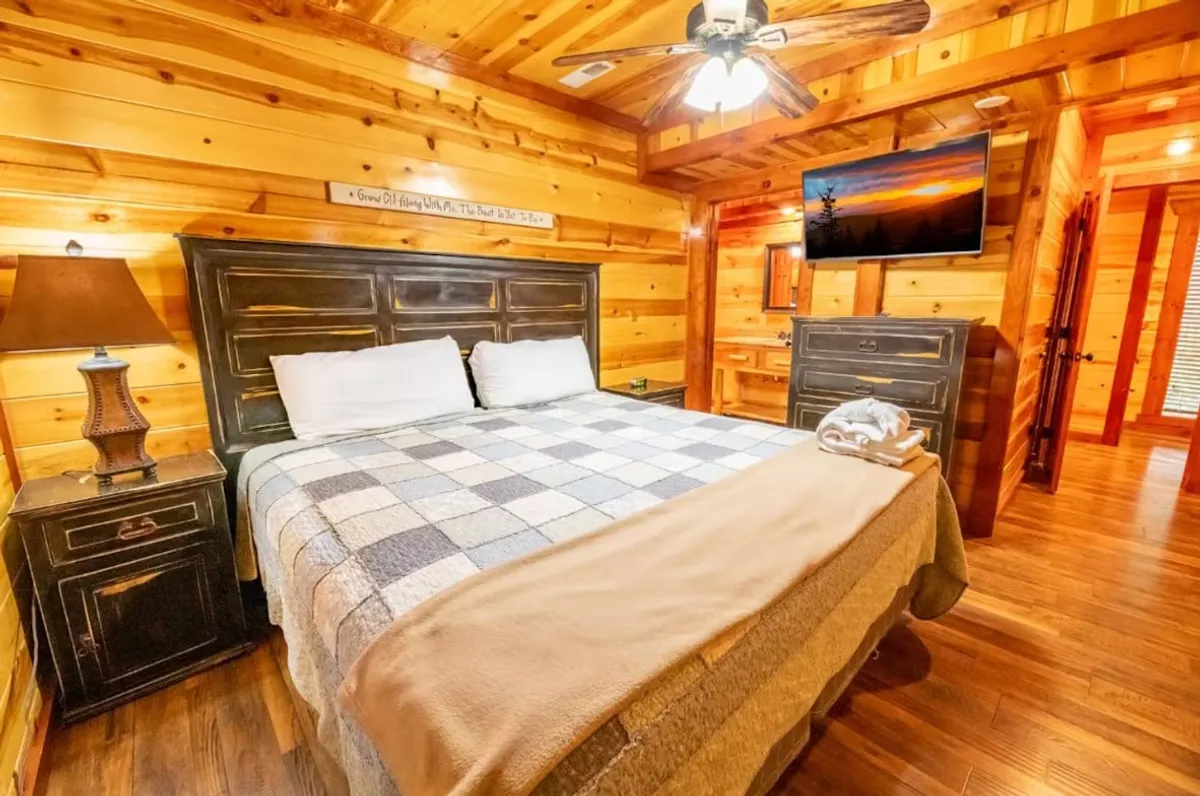 This image showcases a cozy bedroom with wooden walls, a king-sized bed dressed in inviting linens, a nightstand with a lamp, and a wall-mounted TV. The warm lighting and rustic textures create a tranquil, restful atmosphere, ideal for relaxation after a long day.