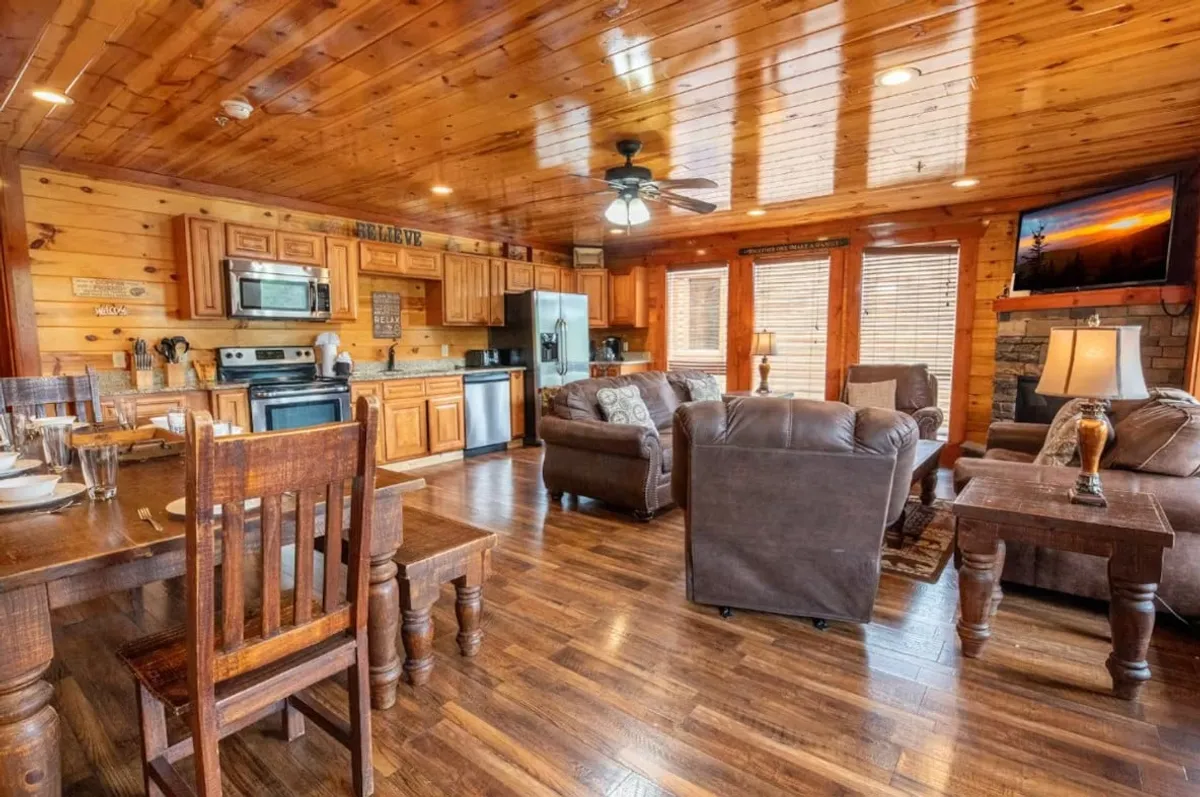 A warm and inviting interior scene featuring a wooden dining table surrounded by comfortable armchairs, a modern kitchen with high-end appliances in the background, and a stone fireplace along one wall. The TV displays a sunset, echoing the scenic beauty outside.
