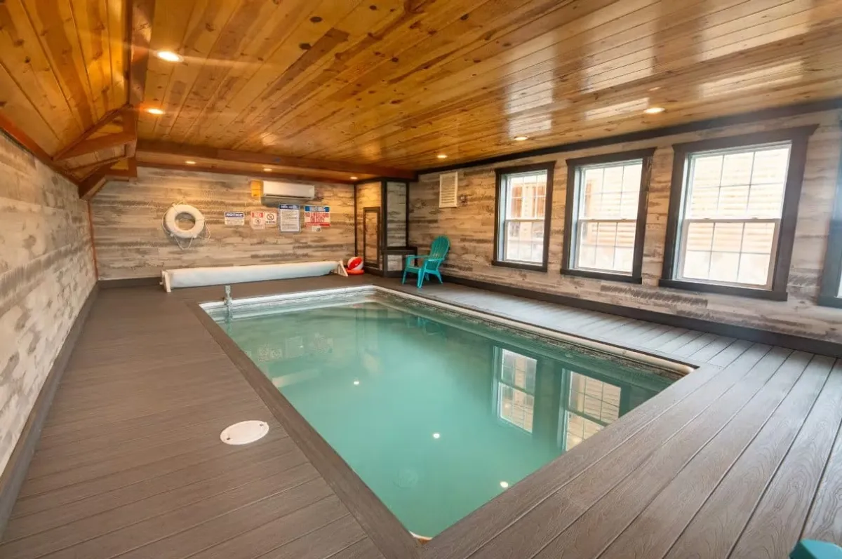 This image showcases a sparkling indoor swimming pool set beneath a wooden ceiling, with large windows allowing natural light to illuminate the tranquil space. A blue chair sits by the pool, inviting relaxation or quiet observation, and the pool's generous size hints at space for both swimming and lounging.
