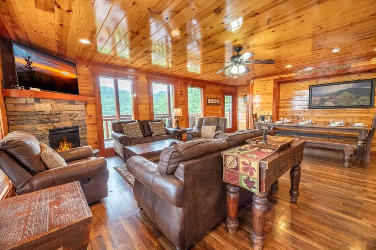 A spacious, open-plan living area with comfortable leather sofas facing a beautiful stone fireplace. Large windows fill the room with light and frame sweeping mountain views. A dining table, set for a meal, sits nearby, seamlessly connecting the living, dining, and kitchen spaces.