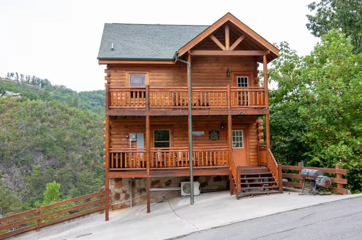 A striking exterior shot of a log cabin with inviting balconies set amidst lush natural surroundings. The image shows a spacious porch ideal for family gatherings or quiet moments, with warm lighting giving the cabin a welcoming glow. The natural landscape frames the scene, providing a strong sense of place.