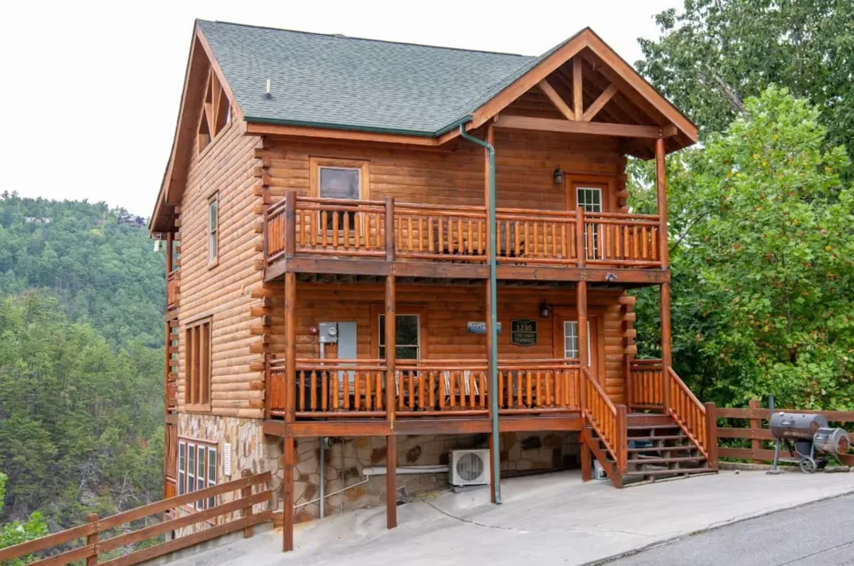 This image displays the log cabin's exterior, featuring spacious balconies and surrounded by lush greenery. The stone foundation and wooden railings evoke rustic charm, while the setting hints at an inviting sanctuary for group gatherings and outdoor activities.