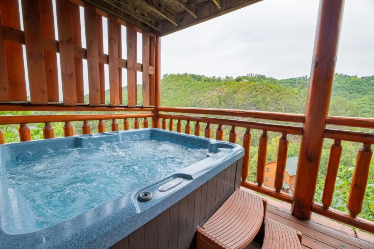 A hot tub is positioned at the edge of a wooden deck, framed by a rustic railing with an expansive view of the surrounding mountains. The scene is tranquil, with golden hour light enhancing the cozy, inviting atmosphere.