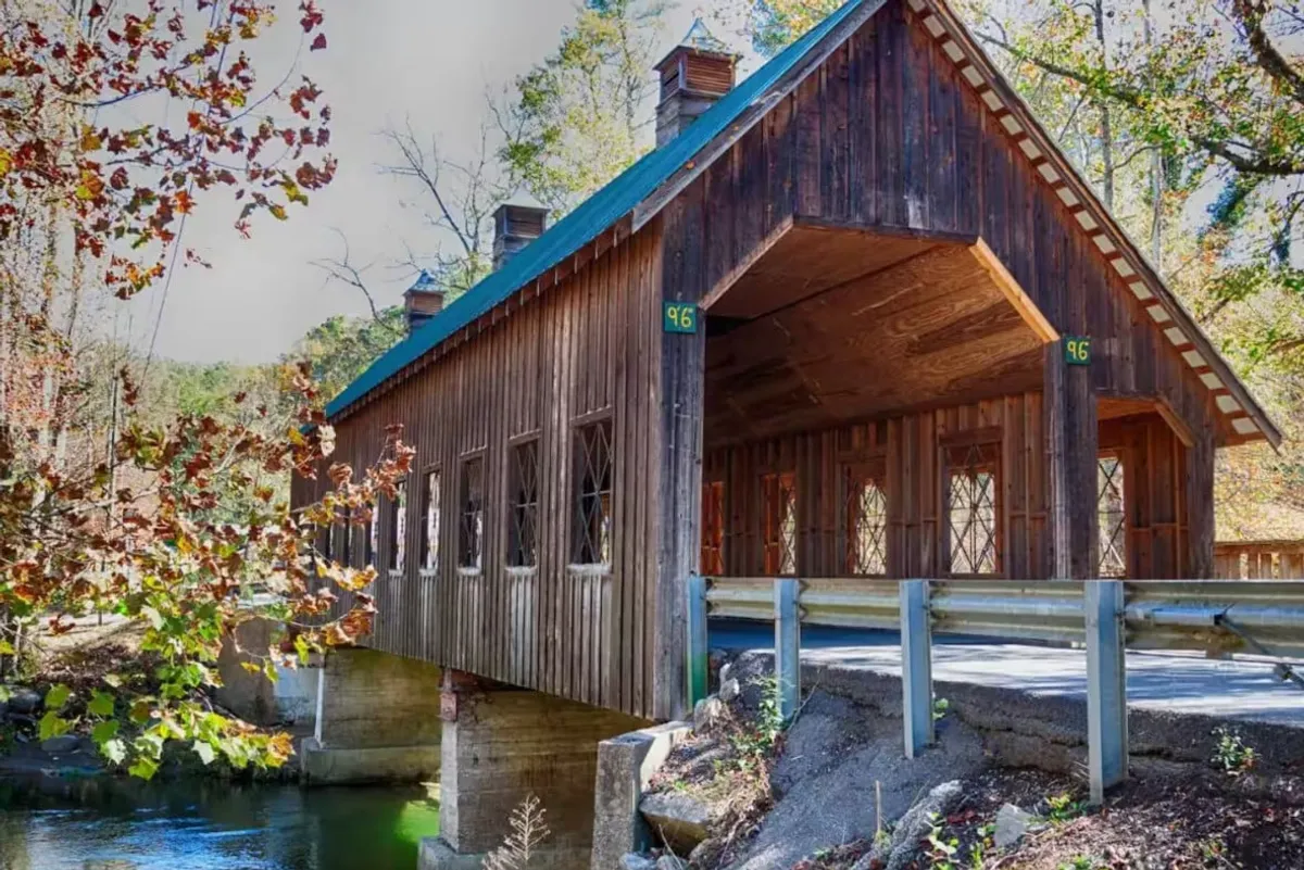 A charming covered bridge stretches over a gentle stream, surrounded by lush trees and reflected in the calm water below. The inviting scene hints at quiet strolls, leisurely exploration, and hidden gems waiting to be discovered nearby.