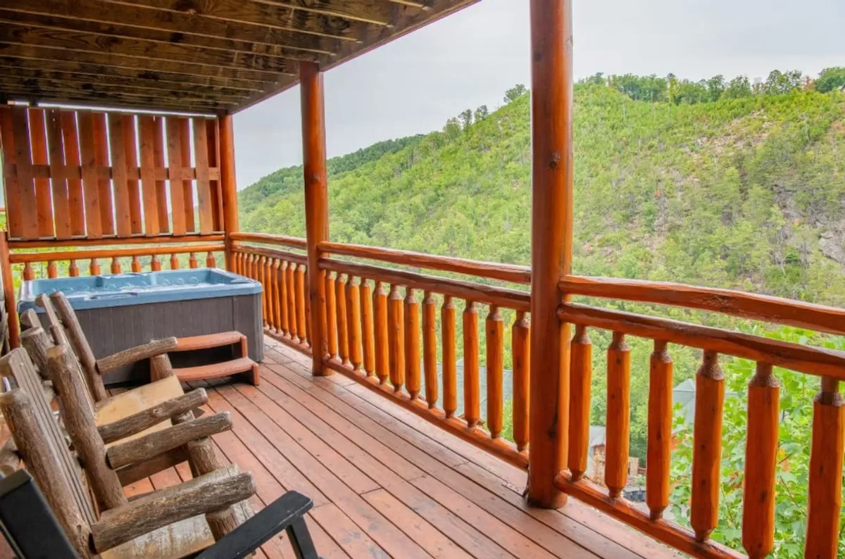 A spacious wooden deck features a bubbling hot tub overlooking sweeping mountain views. Comfortable wooden chairs are arranged near the railing, offering a perfect spot for relaxing or socializing while immersed in nature.