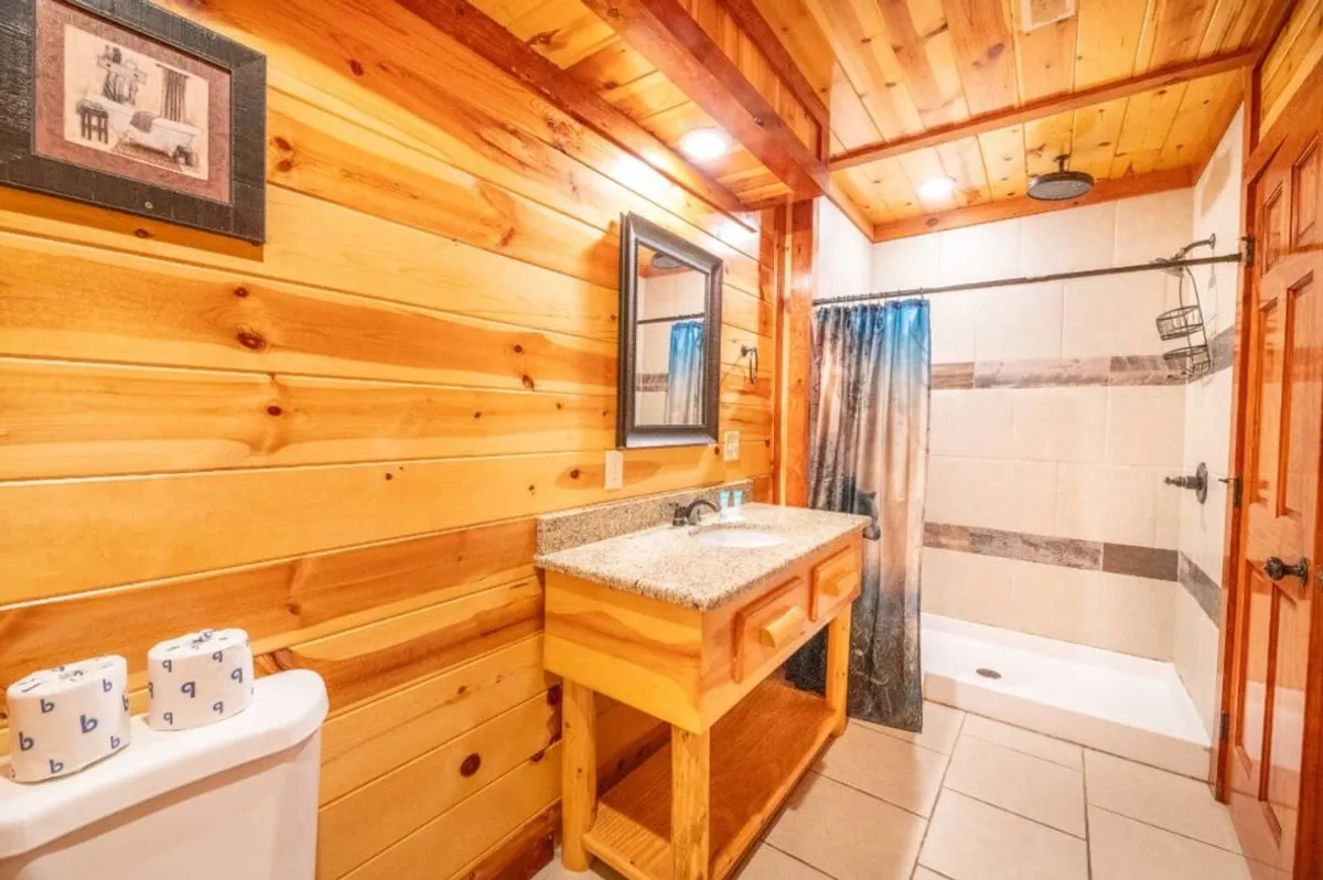 This bathroom features a wooden vanity with granite countertop, a large mirror, a walk-in shower, and warm wooden wall accents. Natural light filters into the space, enhancing its spa-like, inviting atmosphere.