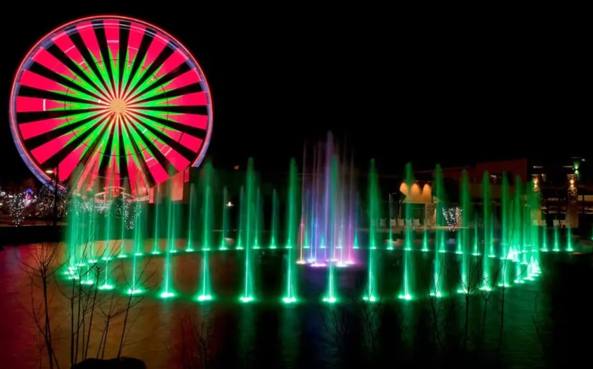 A vibrant nighttime scene featuring a large illuminated Ferris wheel and a colorful water fountain, set against a night sky. The area is lively, with people enjoying the thrill of rides, taking photos, and strolling along the waterfront.